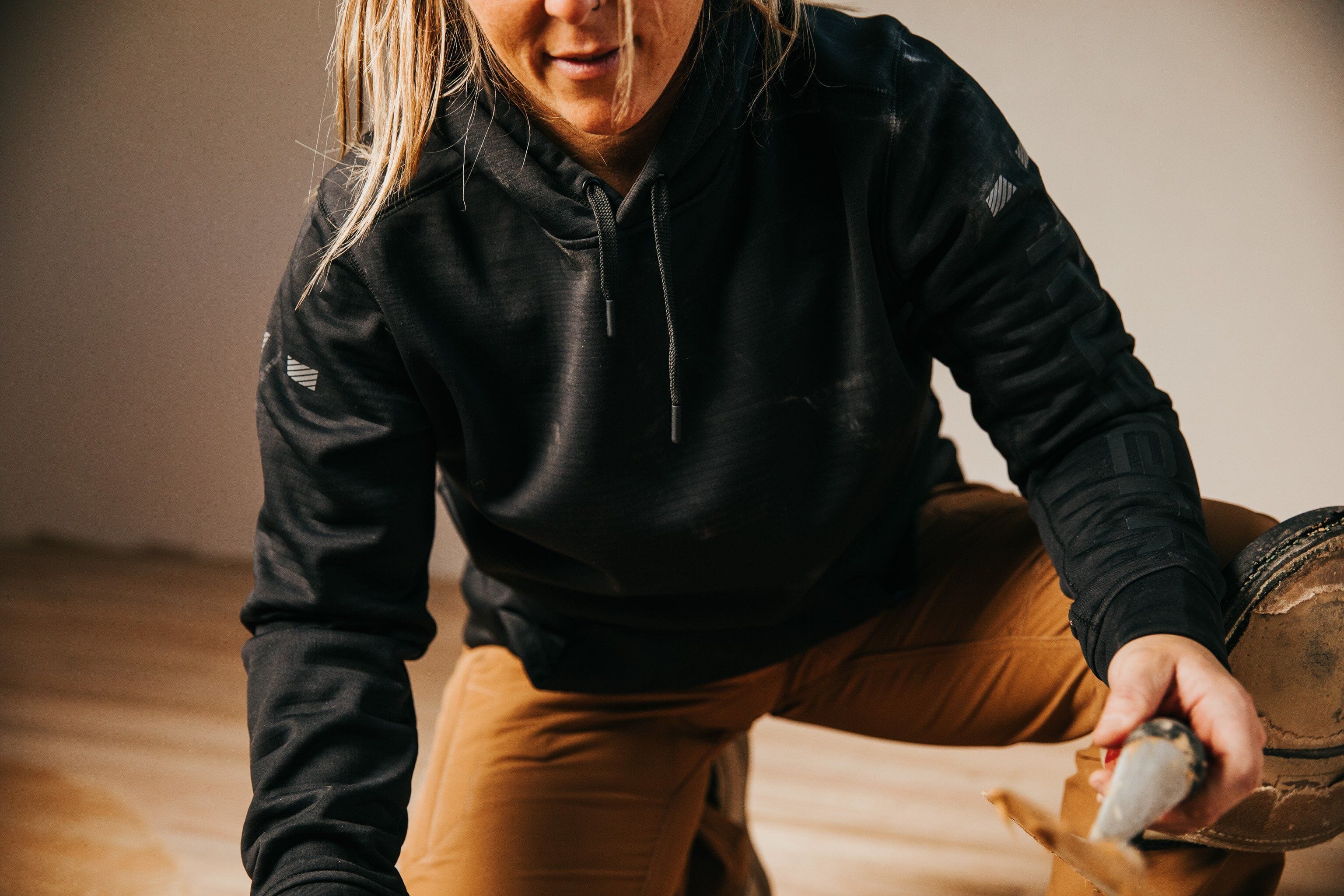 A painter wearing brown T2 Truewerk WerkPants crouches down as she works on the wood floor during a home remodel.