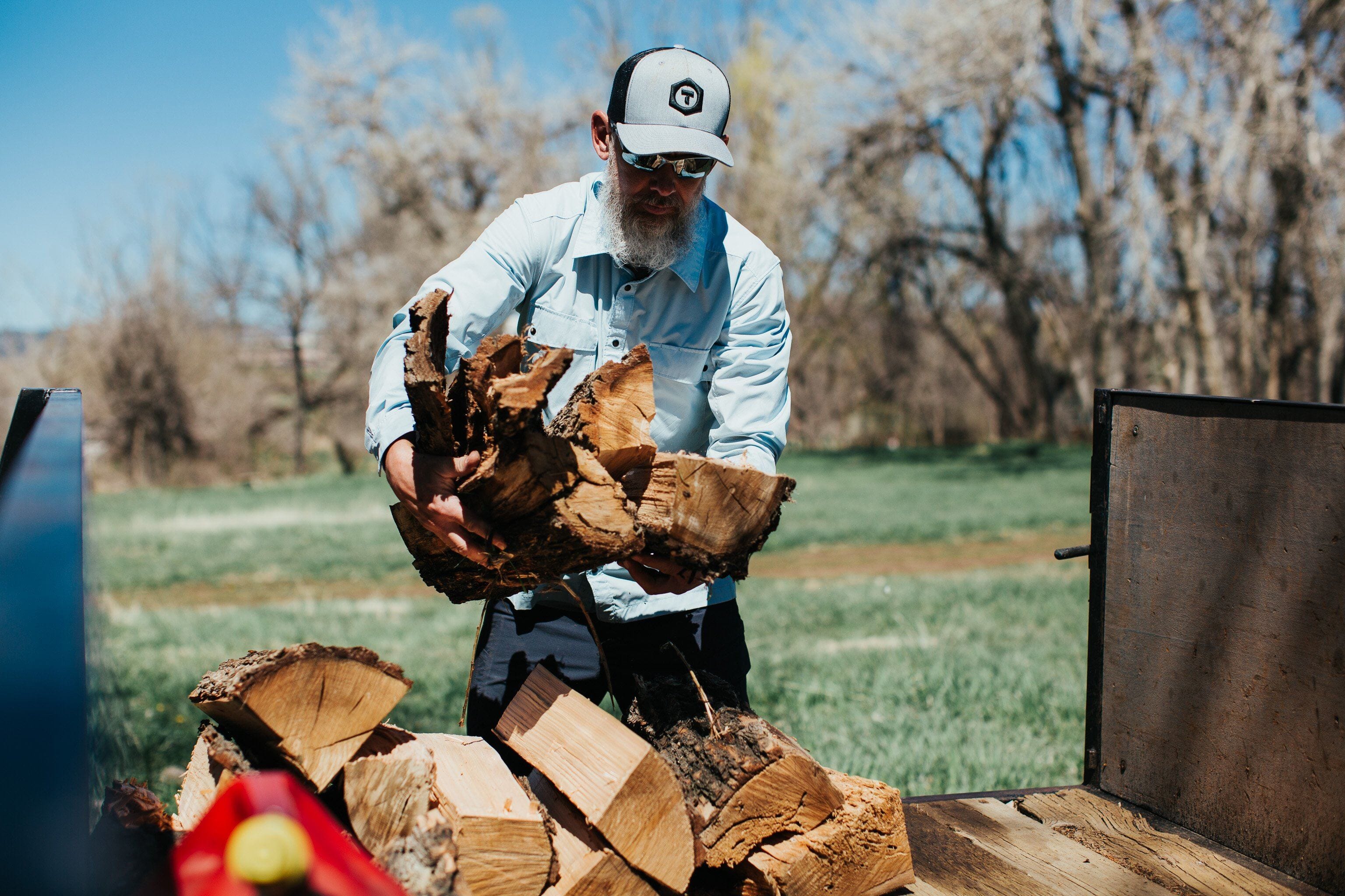 A trade professional in dark blue Truewerk WerkPants, a button shirt, and a Truewerk cap hauls an armful of firewood onto a truck.