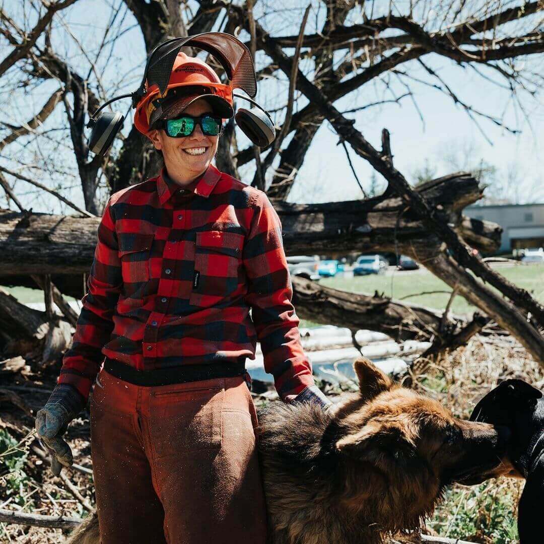 A female arborist stands with her dog in front of a felled tree.