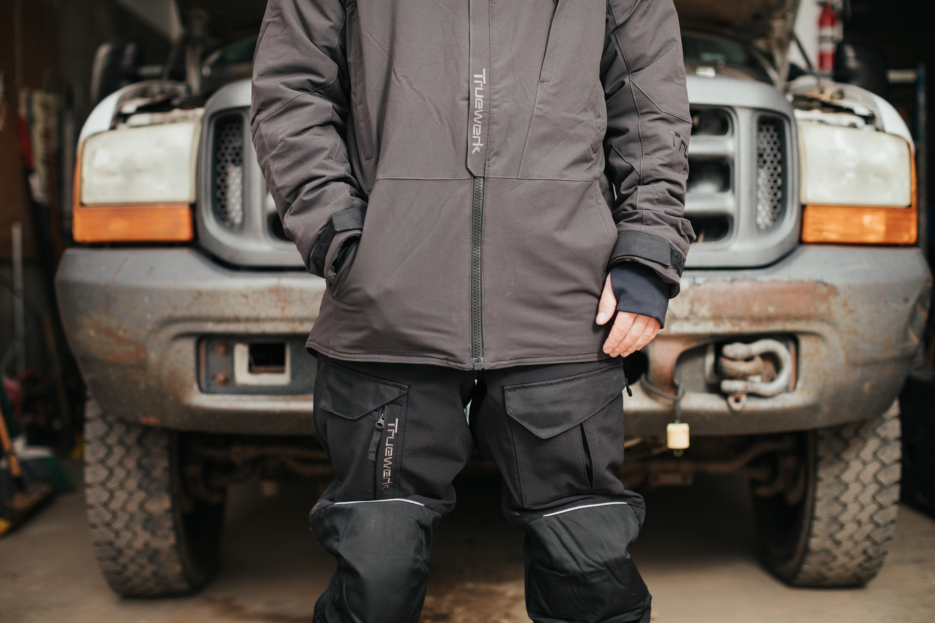 A male auto mechanic wears a gray Truewerk WerkBib and stands in front of an automobile with the hood up.