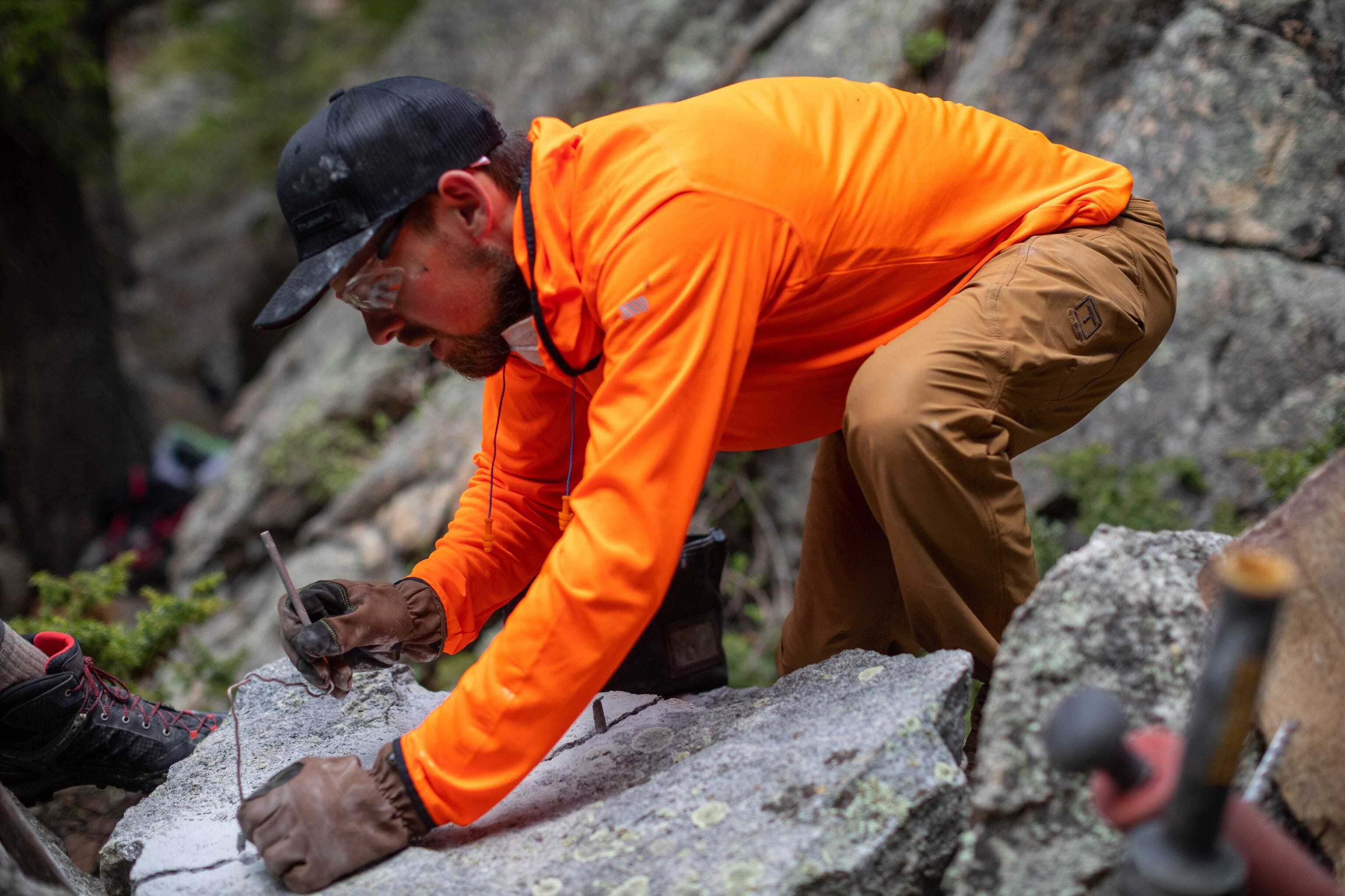 A technician at Little Eden wears orange and tan Truewerk workwear and leans over a boulder as he works on a project.