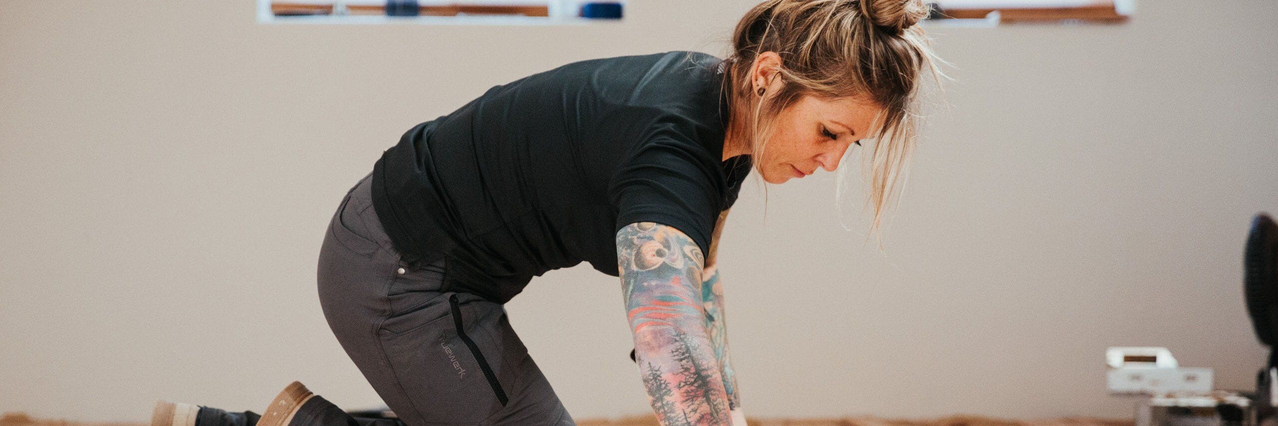 A trade professional with colorful tattoos and dark gray Truewerk workwear pants works on a wood floor during a renovation.