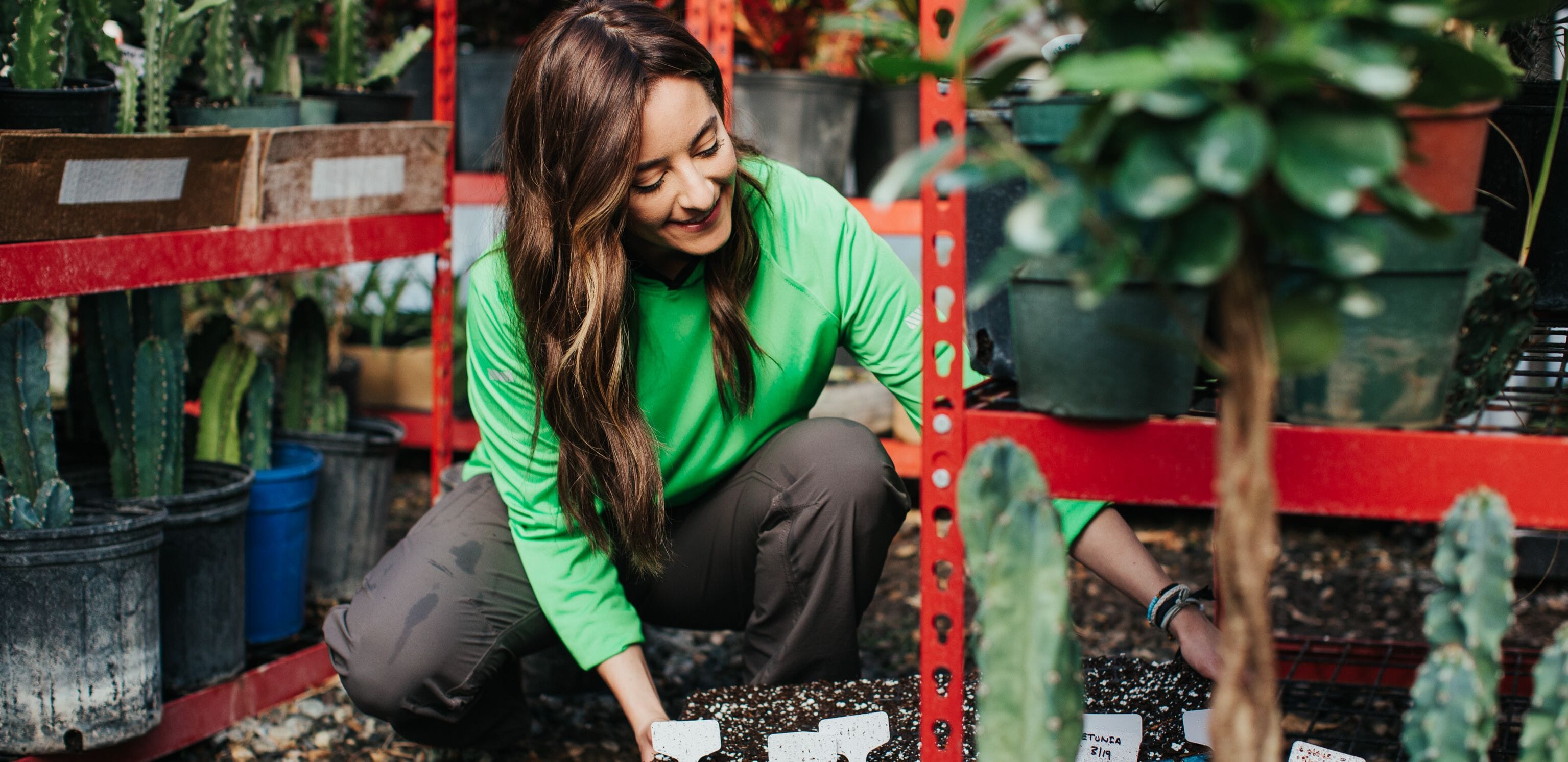 A plant nursery professional wears gray Truewerk WerkPants and a green WerkHoody as she crouches down to tend to the plants.