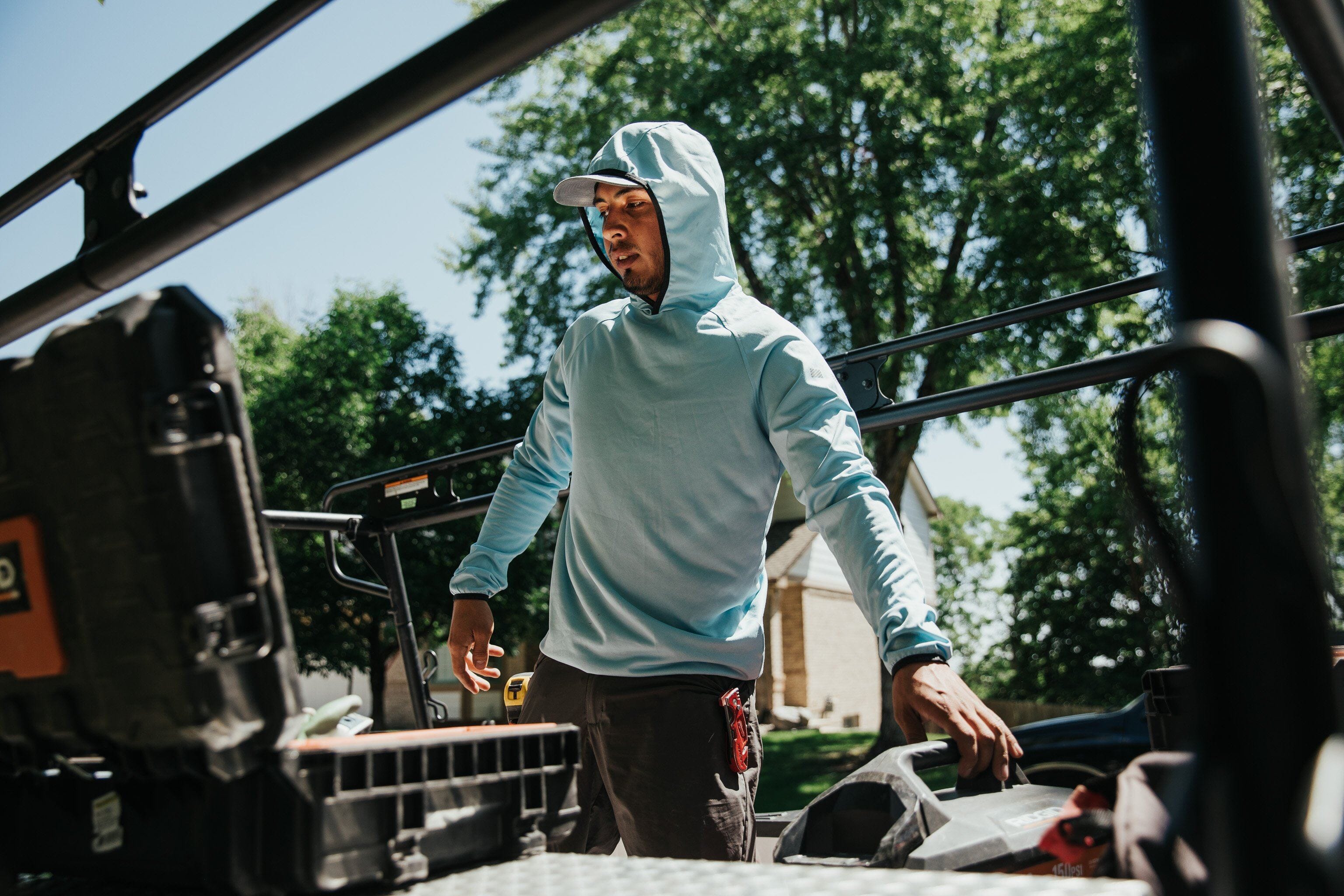 A construction professional wears a blue Truewerk sun hoody over his cap as he works outdoors on a piece of machinery.