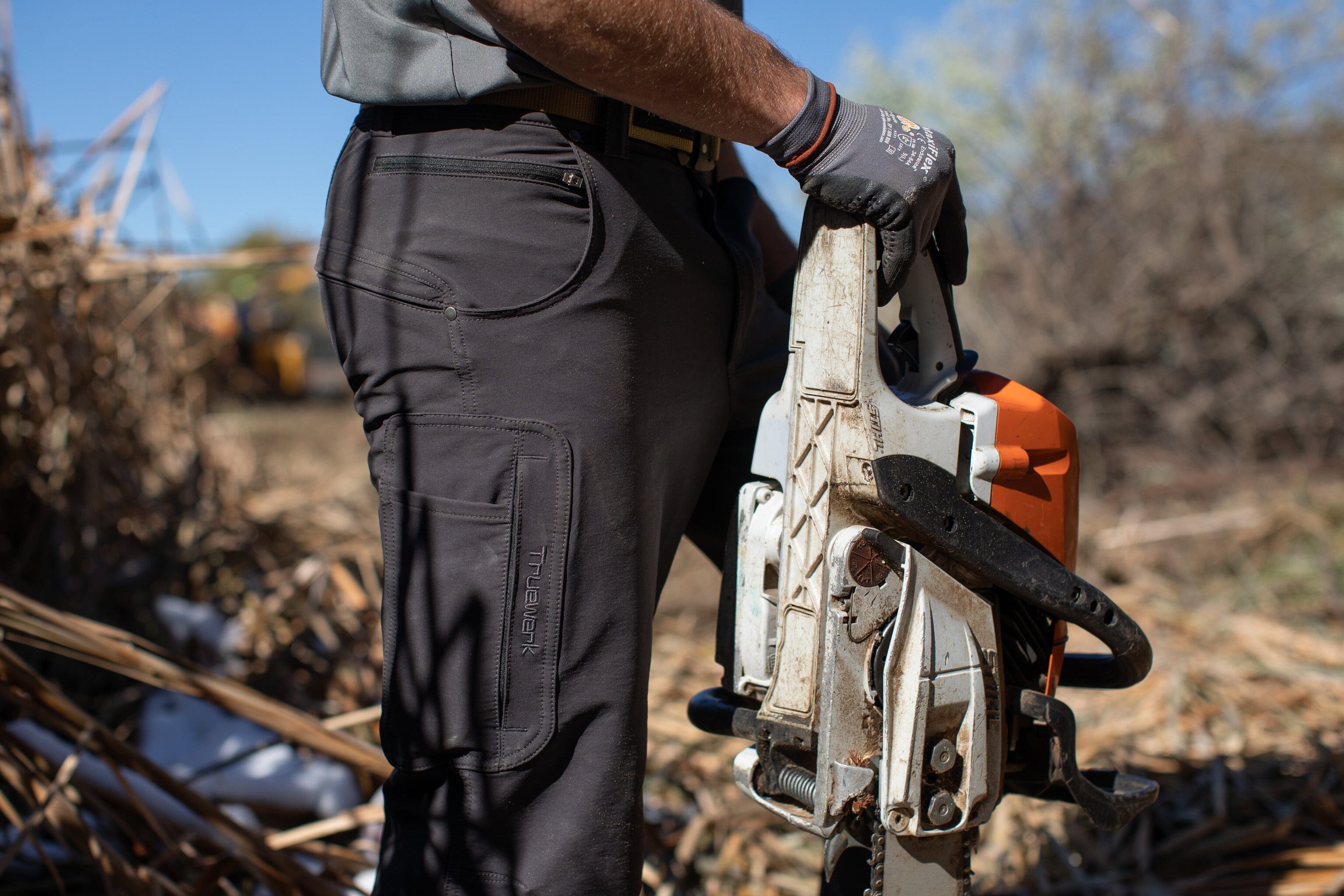 A male trade professional wears dark blue Truewerk WerkPants as he works with a chainsaw to clear brush at a job site.