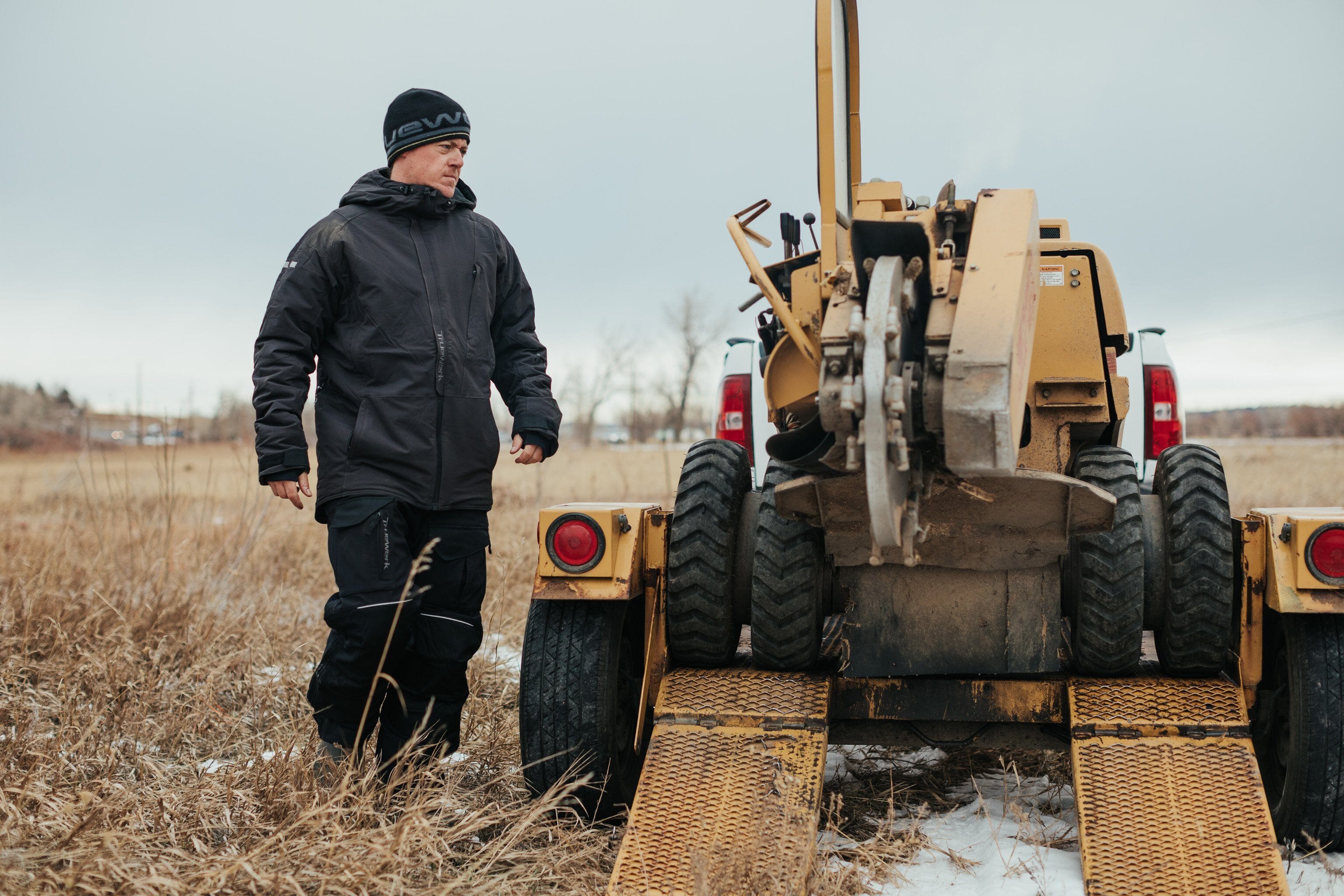 A trade professional wears a Truewerk hat and coat in cold weather as he unloads a piece of machinery from a vehicle.